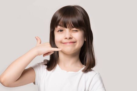 Isolated on beige wall head shot cutie 6 years old girl winking with eye look at camera makes with fingers telephone handset as symbol gesture of call me shaka sign. communication mobile tech concept Stock Photo