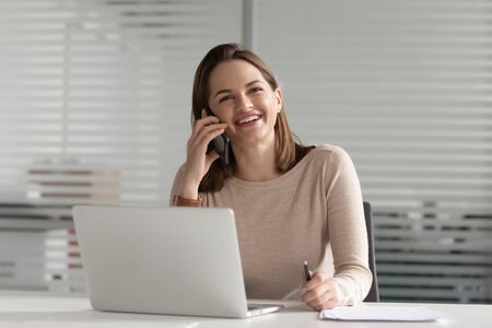 Smiling businesswoman talking on phone make business call at work, happy young entrepreneur female secretary consulting client chatting with friend laughing having mobile conversation in office