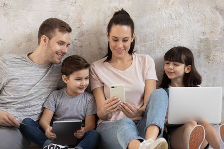 Happy addicted to technology family of four young parents together with little children sitting together on warm heated floor in living room at home, using smartphones, laptop and digital tablet.