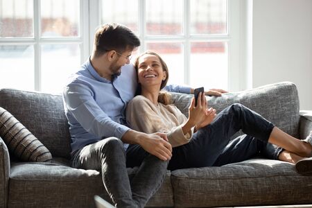 Smiling young man embracing lying on couch happy wife. laughing millennial mixed race woman holding smartphone, showing funny video or photos to husband. positive family couple relaxing on sofa.