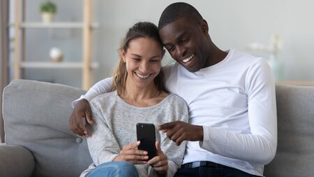 Happy young mixed race family couple sitting on sofa in living room, hugging, bonding, holding mobile phone, using apps, shopping, watching funny movies or pictures, holding video call with friends.