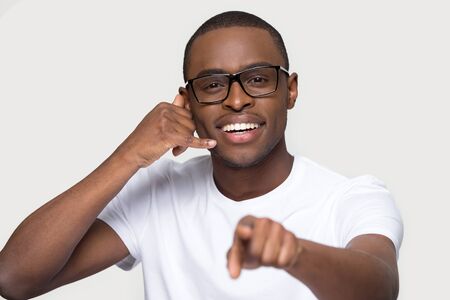 Head shot portrait of smiling african american man in glasses showing call me gesture, looking, pointing finger at camera, happy male with phone shaped hand close up, isolated on studio background