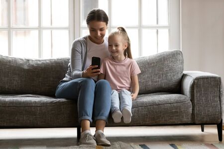 Full length happy mum sitting on cozy sofa with smiling adorable child daughter, looking at smartphone screen. pleasant nanny showing mobile educational application to interested small kid girl.