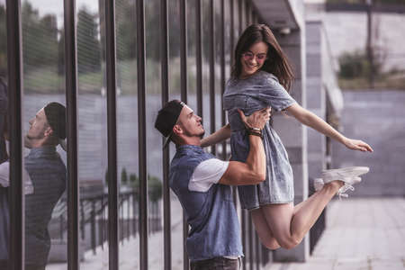 Stylish young couple is having fun and smiling while walking outdoors, guy is lifting his girlfriend - 90996481