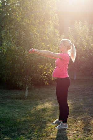 Pregnant woman doing exercises with dumbbells. Expectant female practicing fitness in garden early in morning, sunrise light. Health an body care, healthy pregnancy, sport concept