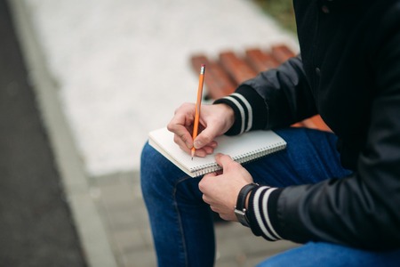 Student in a black jacket sits in a park on a bench writes down his thoughts in a notebook handsome boy