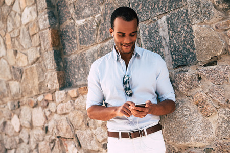 Enjoying his favorite music. Handsome young African man in headphones carrying smart phone and looking at it with smile while standing against the stoned wall outdoors