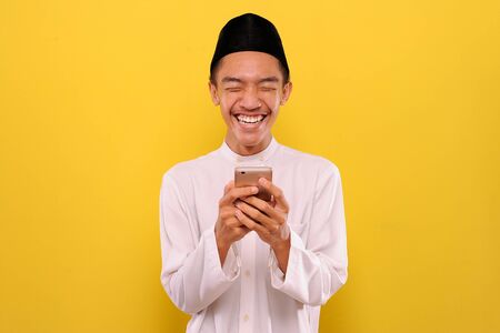 Young asian muslim man read funny messages on smartphone, isolated on yellow background