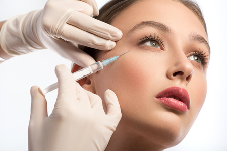 Serine young woman is getting facial botox injection. Beautician hands in gloves holding syringe near her face - 63529440