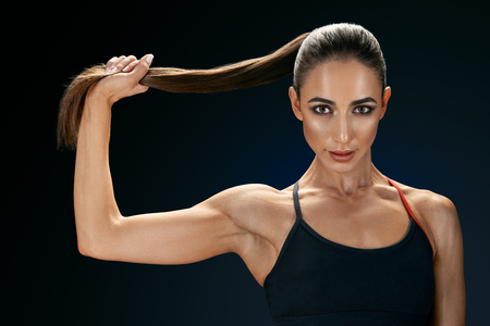 Beautiful young fit woman in sportswear holding her ponytail over black background strong healthy hair clipping path female bodybuilder with power hand confidental looking forward.