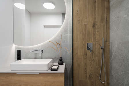 Salle de bains moderne avec carrelage en bois dans la cabine de douche à côté d'un grand miroir rond avec lumière led au-dessus du lavabo