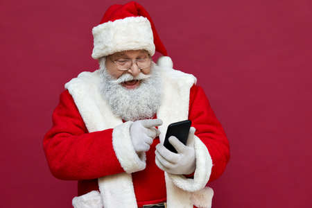 Happy excited santa claus using smart phone isolated on red background.