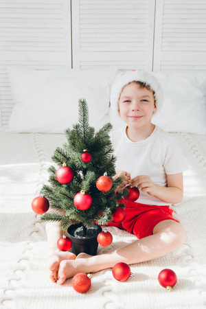 cute boy in red santa hat decorates a small christmas tree balls sitting on the bed - Small Christmas Tree Ornaments cute boy in red santa hat decorates a small christmas tree balls sitting on the bed - Small Christmas Tree Ornaments