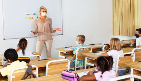 Female teacher in protective mask giving lesson to children Фото со стока