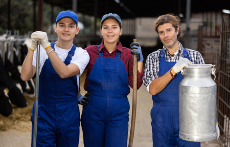 Three dairy farm workers with pitchforks and can of milk stand in cowshed Stock Photo