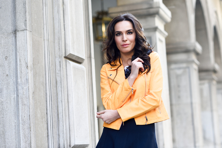 若いブルネットの女性 ファッション モデルは オレンジ色のモダンなジャケットと青いスカートを着ています 長いウェーブのかかった髪型にかなり白人の女の子 都市の背景に女性 の写真素材 画像素材 Image