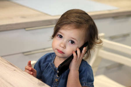 Cute little dark-haired girl 1,5-2,5 in a jeans dress is talking to someone on a smartphone in the kitchen.