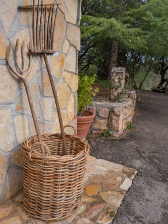Pair of rakes placed in a wicker basket and supported by a rustic wall, with a garden and a fountain in the background Stock Photo