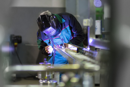 Industrial worker with protective mask welding inox elements in steel structures manufacture workshop. Фото со стока