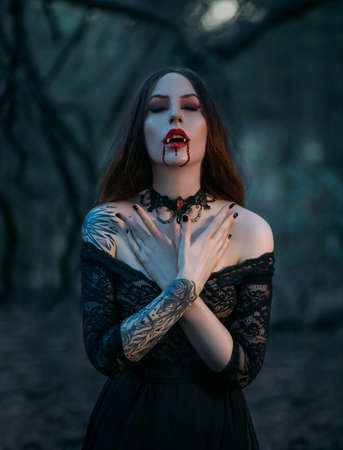 Attractive vampire woman with sharp teeth fangs drops of blood flowing on red lips. close-up portrait of beautiful mouth. festive art make-up. gothic lace choker on the neck. medieval girl Фото со стока