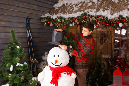 stock photo young white man in winter outfit putting a black hat on indoor snowman with red scarf at wooden house surrounded by christmas ornaments - Indoor Snowman Christmas Decorations stock photo young white man in winter outfit putting a black hat on indoor snowman with red scarf at wooden house surrounded by christmas ornaments - Indoor Snowman Christmas Decorations