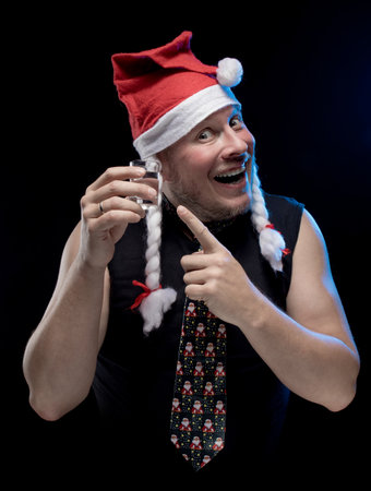 Comic actor man in cap with braids with a glass of vodka on a black background, in anticipation of Christmas and New Year
