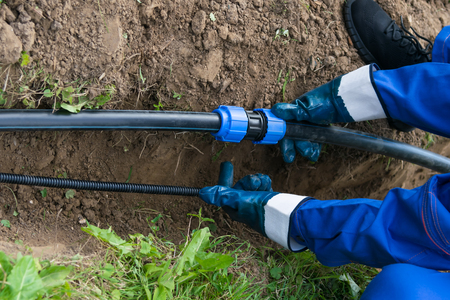 worker's hands hold a black pipe over a dug hole in the ground - 111946824