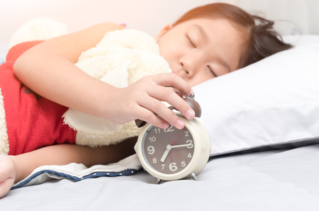 little hand girl reaching to turn off alarm clock on the bed in the morning. Stock Photo