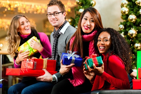 Diversity group of four people - Caucasian, black and Asian - sitting with Christmas presents and bags in a shopping mall in front of a Christmas tree with baubles - 10428072