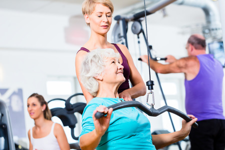 Senior woman doing back training with trainer in gym at machine Фото со стока