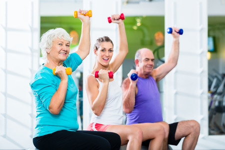 Senior man and women with fitness trainer in gym lifting barbells as sport exercise Фото со стока