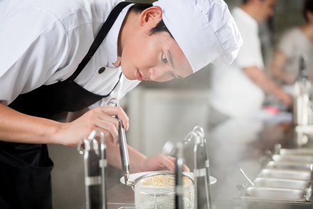 Young Asian chef plating food in a restaurant carefully pipetting garnish onto the side of a plate Stockfoto