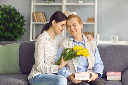 Happy senior woman profitant de cadeaux sur les mères ou la journée internationale de la femme. une jeune fille aimante donne des fleurs et des cadeaux à la vieille maman. petite-fille souriante assise sur un canapé avec grand-mère et la serrant dans ses bras