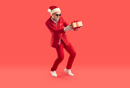 Funny guy veut laisser la surprise sous le sapin de noël. homme sournois en costume rouge, casquette de père noël et lunettes de soleil tenant une boîte-cadeau avec un cadeau secret et marchant sur la pointe des pieds sur fond de studio rouge Banque d'images