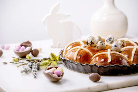 Petits pains chauds traditionnels de pâques faits maison dans un plat en céramique avec des branches de saule en fleurs, des bonbons au chocolat et des œufs de caille sur une table en marbre blanc. cuisine et décoration des fêtes de pâques
