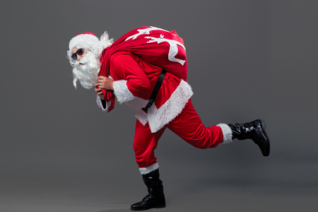 Santa claus with sunglasses runs with the bag in christmas gifts on his back on the white wall.