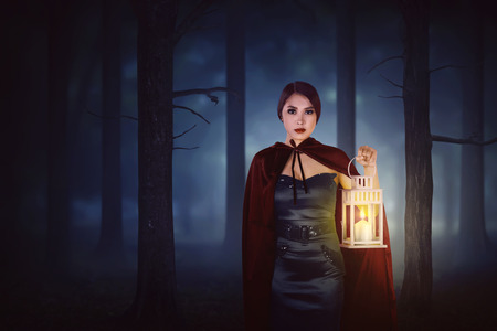 Moonlight lanterns: Young asian witch woman with red cloak walking in forest with a lantern at midnight