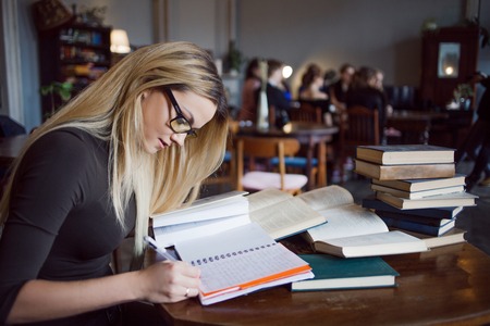 Blonde girl in the library, makes a summary of the textbooks. education, exam preparation. sitting at table surrounded by large number of textbooks