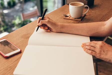 Cropped shot of blogger making notes in notebook at table with cup of coffee in coffee shop