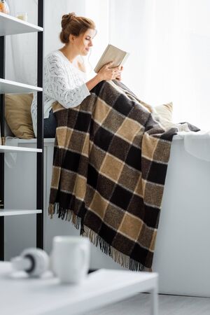 Attractive woman in white sweater with blanket reading book