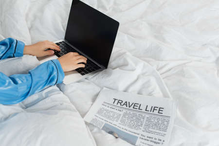 Cropped view of young woman in pajamas using laptop near newspaper on bed