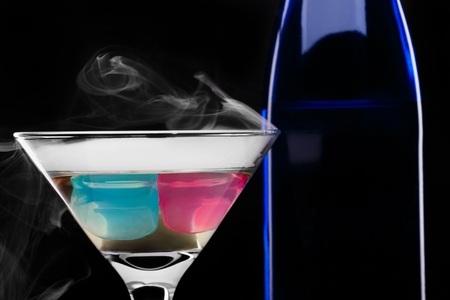 Wine glass with colored ice cubes and dark blue bottle in floated ice vapor on black background