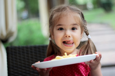 Beautiful laughing little girl sitting at table and holding a plate with French fries, food and drink concept, outdoor portrait - 60414091