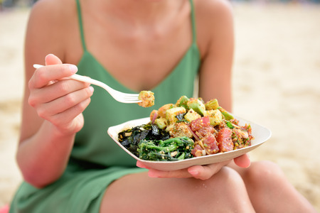 Poke Schüssel Salatplatte. Ein traditionelles lokales Hawaii-Speisegericht mit rohen marinierten Ahi Yellowfin-Thunfischen. Frau, die auf dem Strand isst Mittagessen sitzt. Foto auf Lager