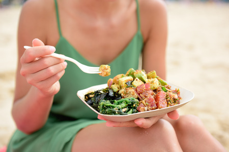 戳碗沙拉盘。传统的夏威夷当地food dish with raw marinated ahi yellowfin tuna fish. Woman sitting on beach eating lunch. Stock Photo