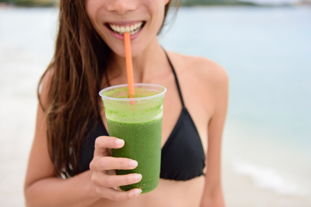 Smoothie vert. Femme tenant le jus de detox de légume vert à l'extérieur en bikini au soleil d'été sur la plage. Mode de vie sain avec belle race mixte modèle féminin asiatique caucasien prendre un régime de nettoyage.