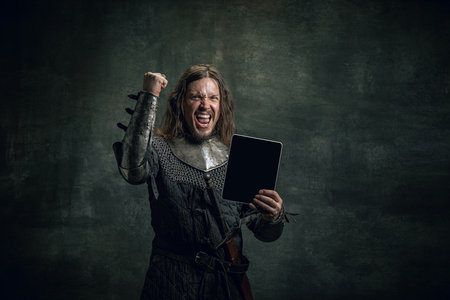 Portret w stylu vintage brutalnego uśmiechniętego mężczyzny, średniowiecznego wojownika lub rycerza z brudną ranną twarzą za pomocą nowoczesnego gadżetu izolowanego na ciemnym tle. porównanie epok