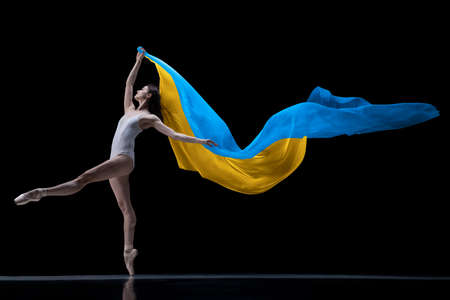Young graceful classic ballerina dancing with cloth painted in blue and yellow colors of ukraine flag on dark studio background. art, peace, freedom and rights