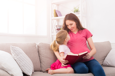 Little girl and her mom reading book sitting on the sofa at home. Mother and daughter study fairy tales. Motherhood, joint activities and interests, early development, caring concept, copy space