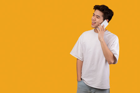 Communication, phone call. portrait of casual smiling asian teen in white t-shirt talking on his cellphone, looking aside at free space, standing and posing isolated over orange studio background